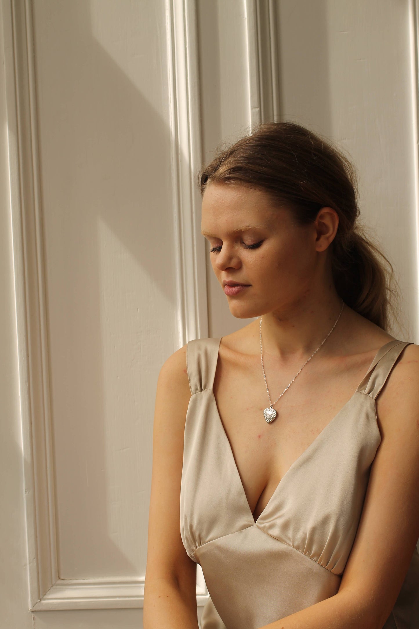 Woman wearing a beige dress with a necklace against a neutral wall.
