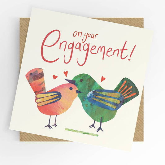 An engagement greeting card with a whimsical illustration of two birds facing each other under the red script text 'on your Engagement!'. The card is shown at an angle resting on top of a brown kraft envelope against a clean white background.