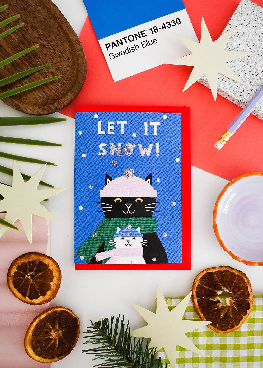 A square greeting card with a blue background and the text 'LET IT SNOW!' featuring a black cat in a pink hat and green scarf holding a white cat. The card is styled in a flat lay with dried orange slices, cream star cutouts, a pencil, a Pantone card labeled 'Sweden Blue', and greenery on a white surface.