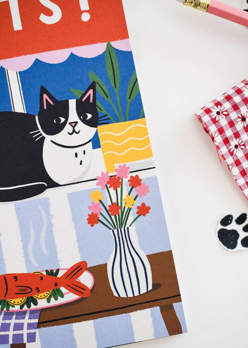 A close-up of a colorful printed card or notebook page featuring a black-and-white cat, flowers in a striped vase, a red fish, and a blue-and-white striped background, photographed on a white surface with a pencil and small accessories partially visible at the edges.