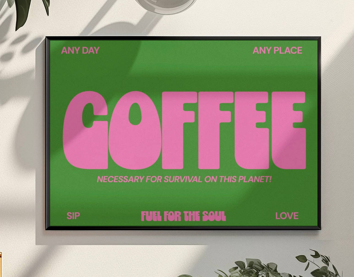 Framed poster with pink 'COFFEE' text on a green background, placed on a light surface with plants around.