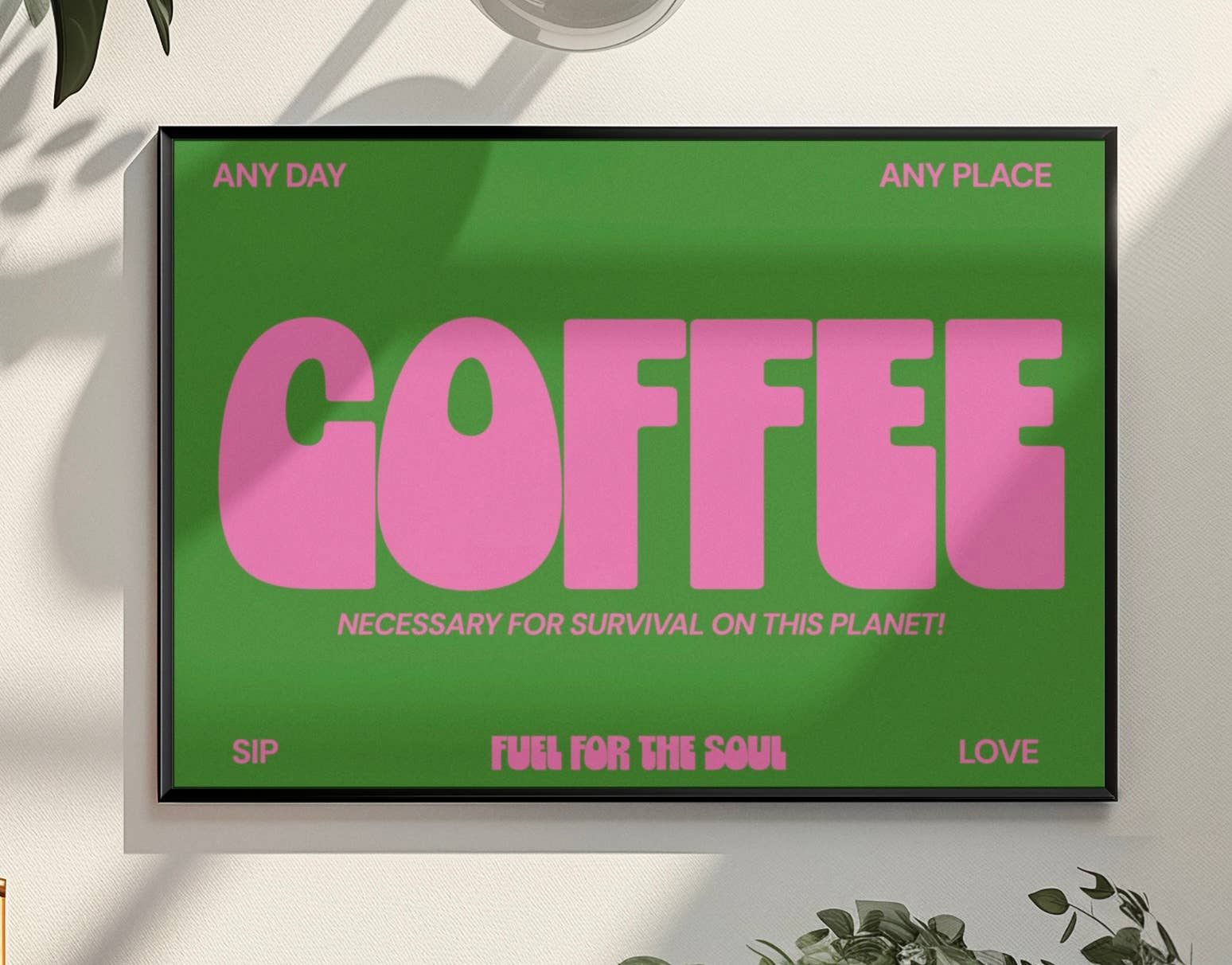 Framed poster with pink 'COFFEE' text on a green background, placed on a light surface with plants around.