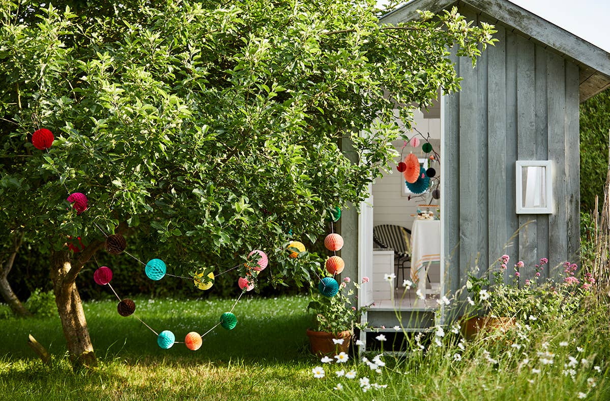 Small wooden cabin in a garden with colorful pom-pom decorations and greenery.