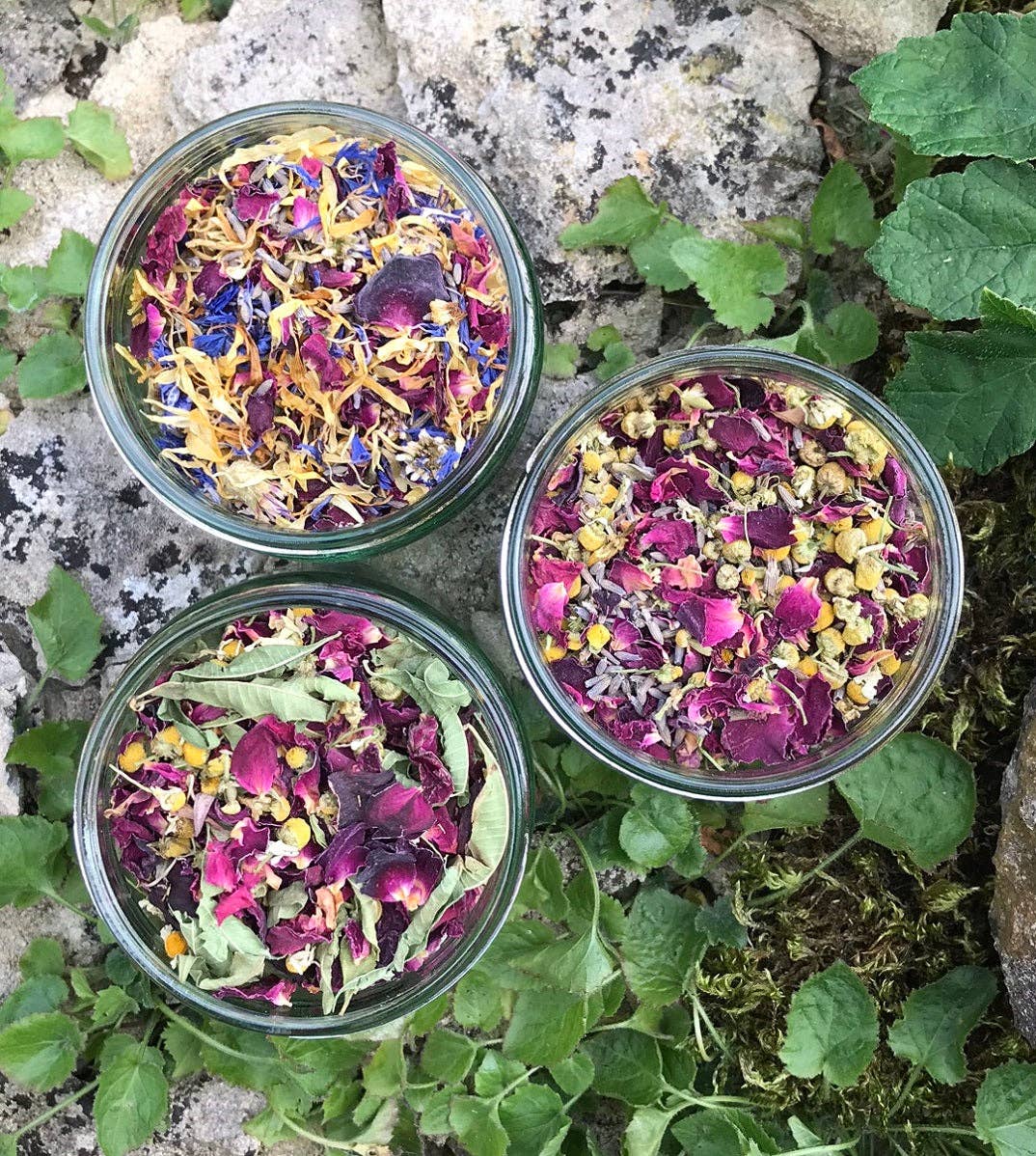 Three small glass jars filled with colorful dried flowers on a natural stone and leaf background.