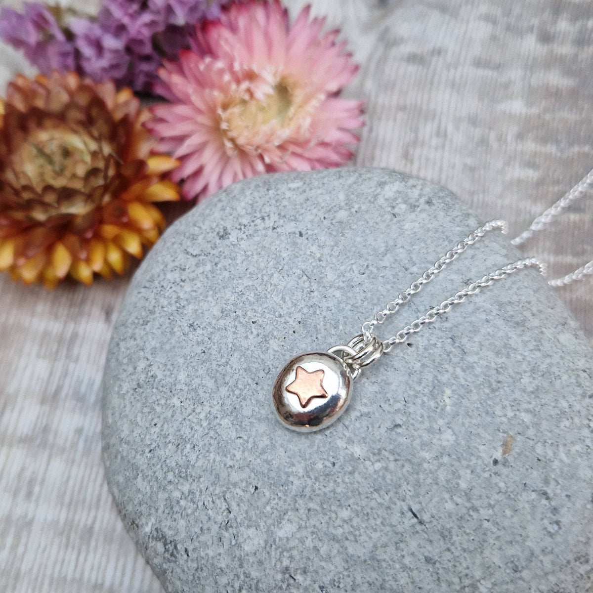 A silver necklace with a small round pendant featuring a star design, displayed on a gray stone with dried flowers in the background.