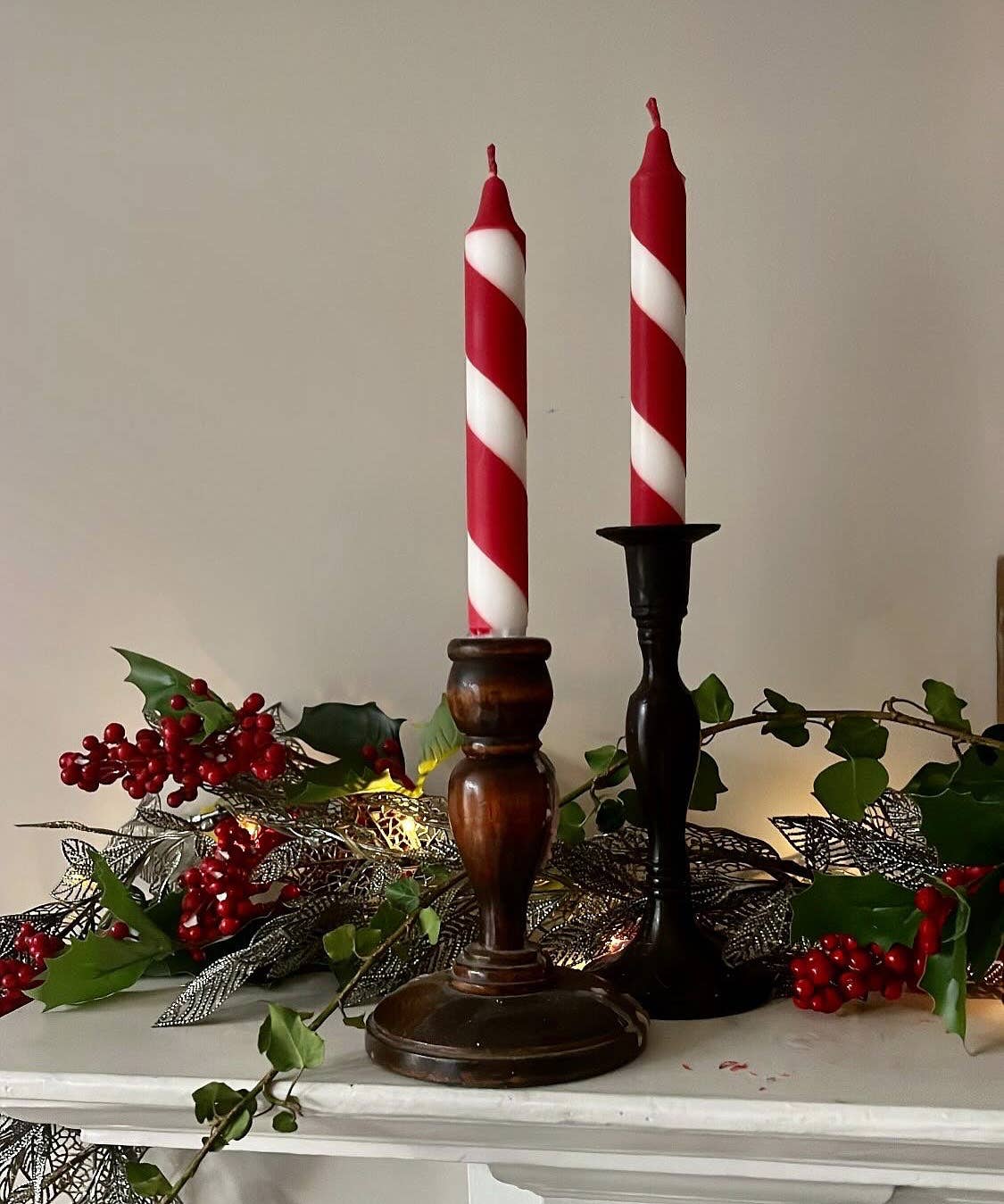 Two red and white striped candles in wooden candlesticks on a decorative surface with greenery.