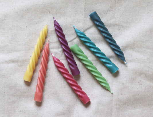 Six twisted taper candles in yellow, coral pink, purple, green, teal, and dark blue, arranged diagonally on a textured beige fabric background.