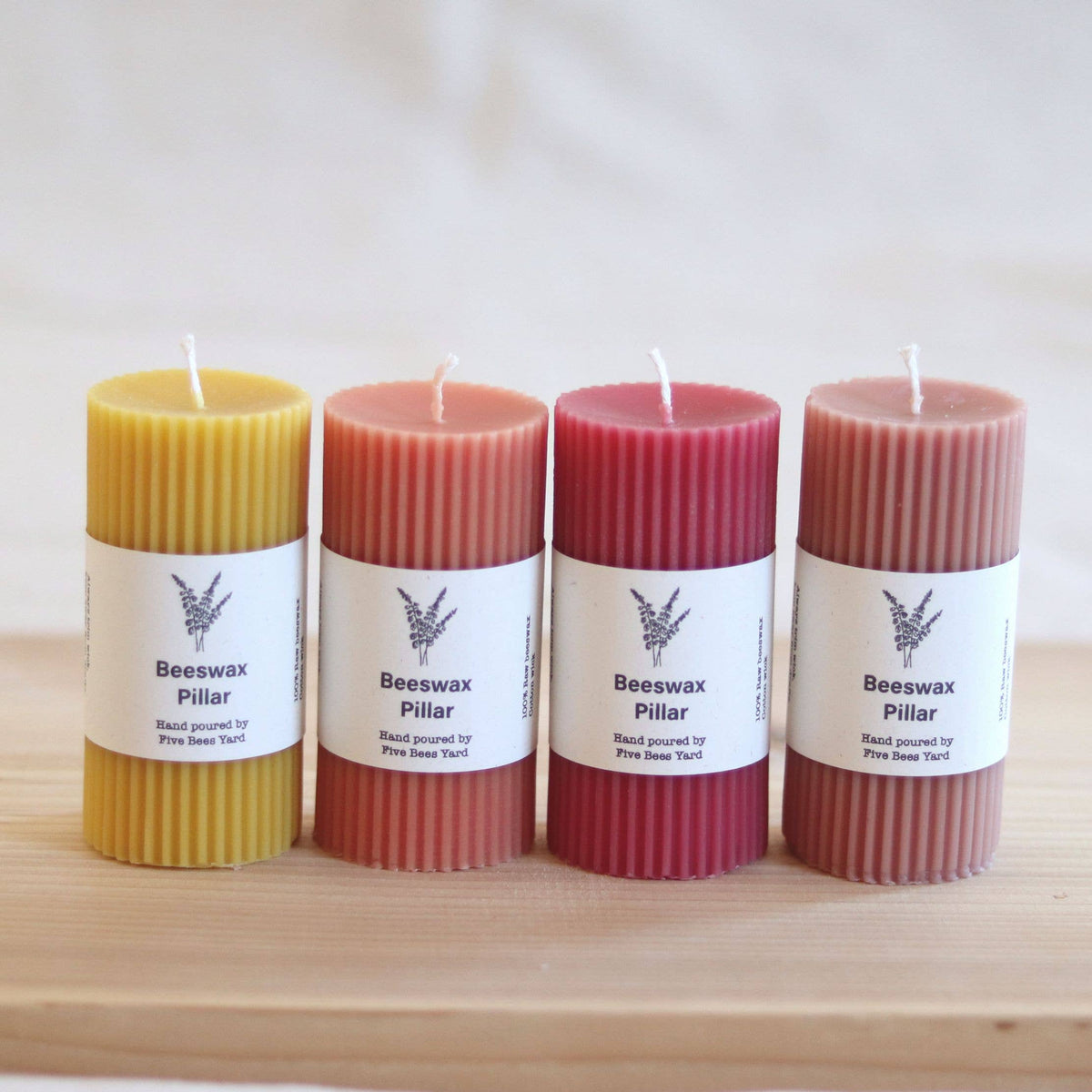 Four beeswax pillar candles in different colors (yellow, peach, dark pink, and mauve) arranged on a light wooden board against a soft, neutral background.