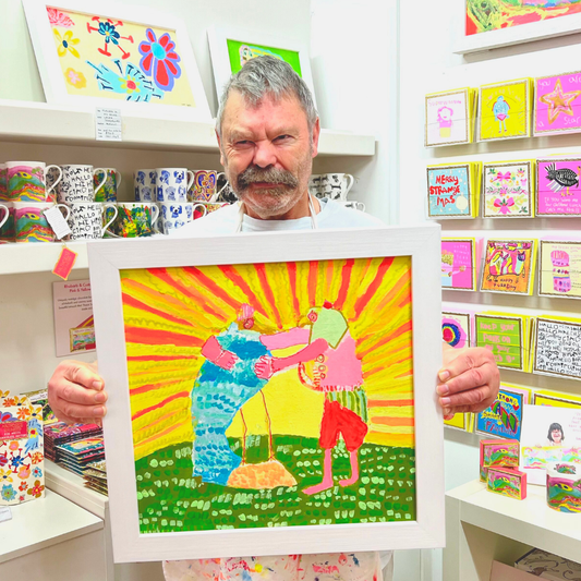 Man holding a framed colorful artwork in a store with shelves of products.