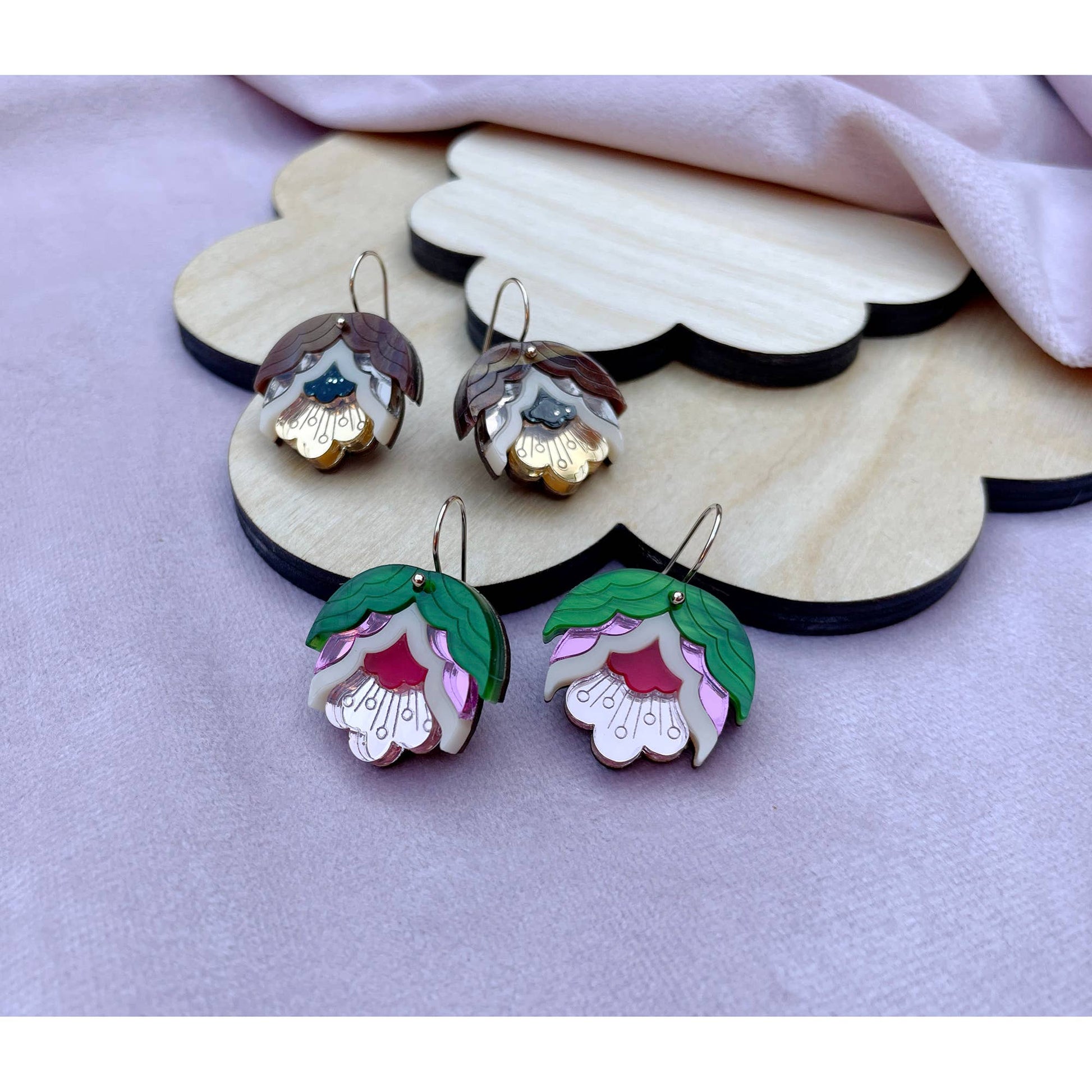 Four flower-shaped earrings displayed on a scalloped wooden stand over a soft lavender fabric background. Two earrings are shown from the front with green leaves, pink inner petals, white outer petals, and gold hooks, while the other two are angled to show the back with brown and gold detailing.