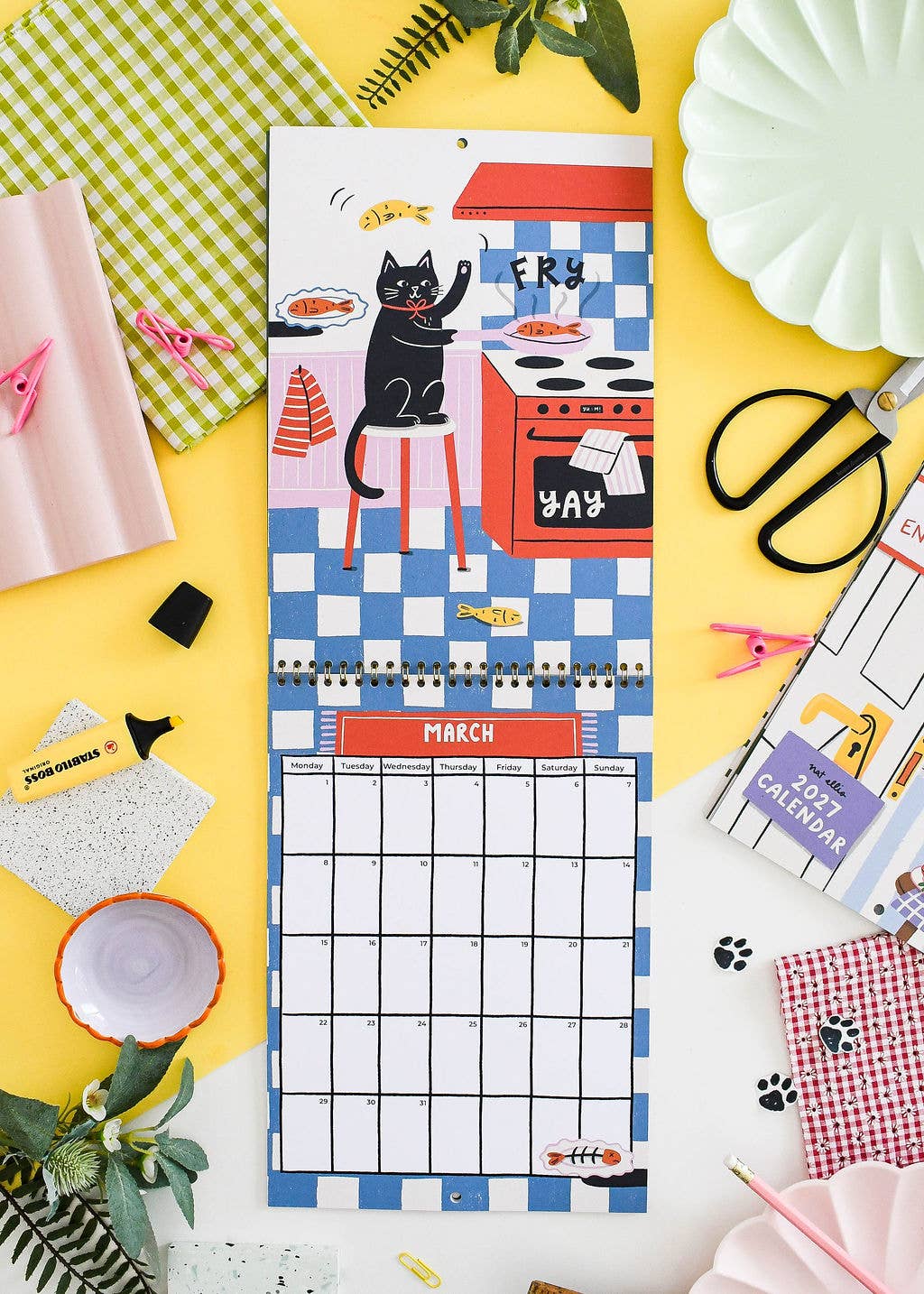 A colorful monthly calendar page featuring a black cat cooking at a stove, laid flat on a bright yellow surface surrounded by styled craft and kitchen props.