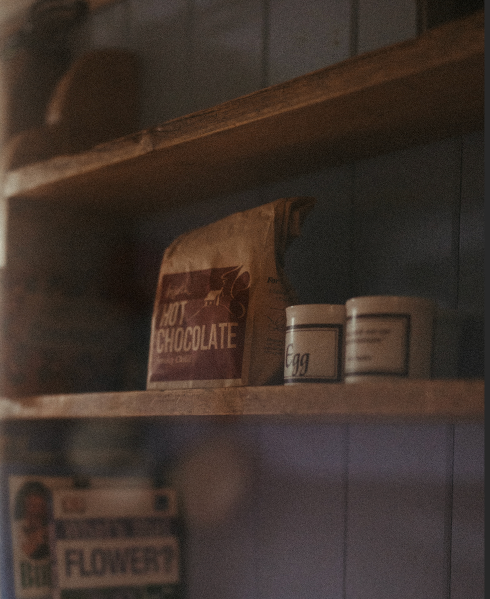 Shelves with a bag of hot chocolate, a jar labeled 'Egg', and a box of 'Flowers' on a tiled wall background.