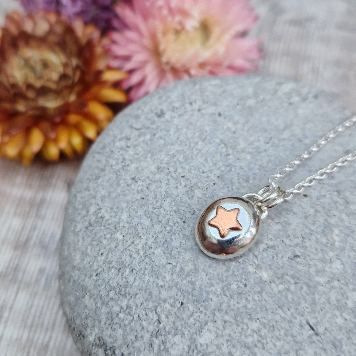 A silver necklace with a small oval pendant featuring a raised rose-gold star, displayed on a smooth gray stone with dried flowers in the background.