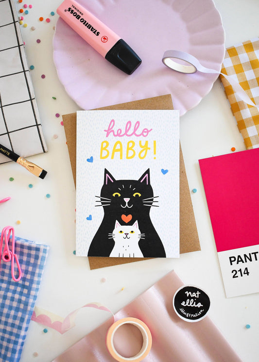 Greeting card with a black cat illustration and 'hello BABY!' text, surrounded by stationery items on a white surface.