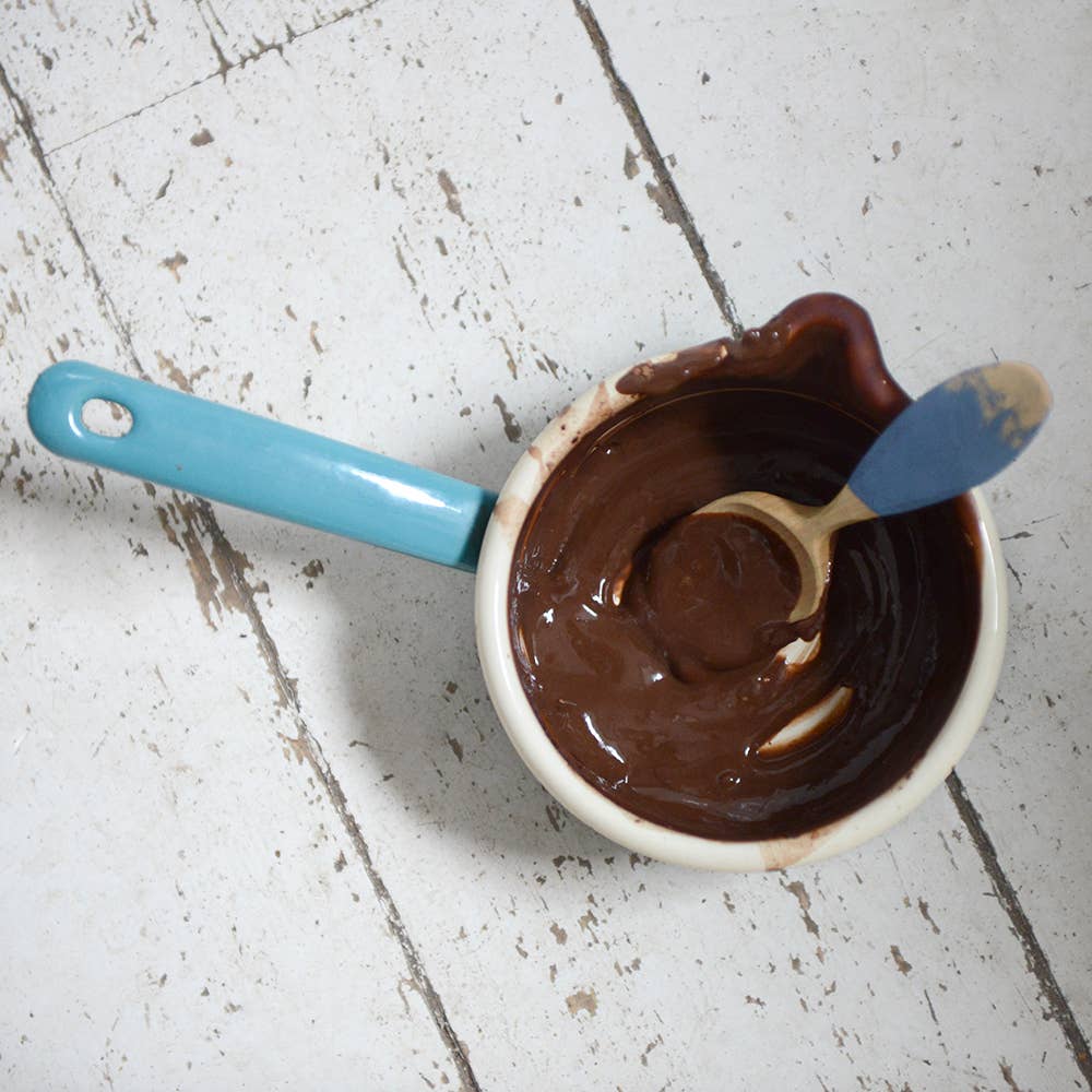 Cup of melted chocolate with a blue-handled spoon on a white wooden surface