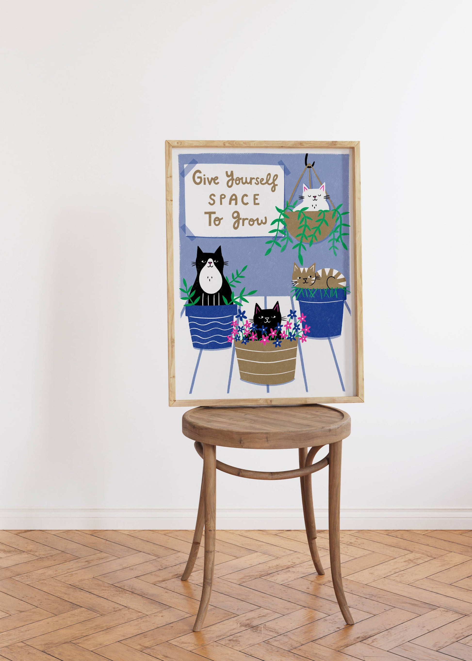 Framed artwork with cats and plants on a wooden stool against a white wall.
