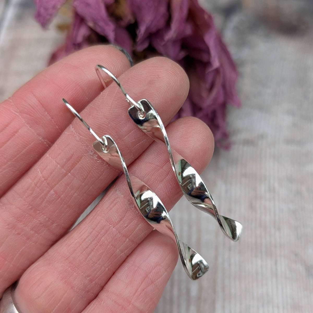 A pair of silver dangling earrings with a twisted spiral design, held in a person's hand against a soft, blurred background with dried purple flowers.