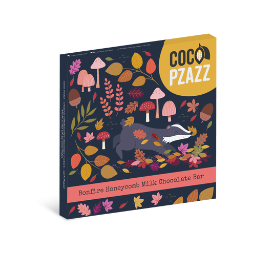 A boxed chocolate bar package on a plain white background. The front shows a dark navy forest illustration with autumn leaves, mushrooms, acorns, and a gray-and-white fox, plus a yellow label reading COCO PZAZZ and a pink band reading Bonfire Honeycomb Milk Chocolate Bar.