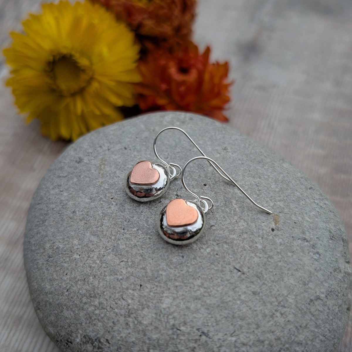 A pair of small heart-shaped dangle earrings with shiny silver-toned metal and pale pink heart centers, resting on a smooth gray stone with yellow and orange flowers blurred in the background.