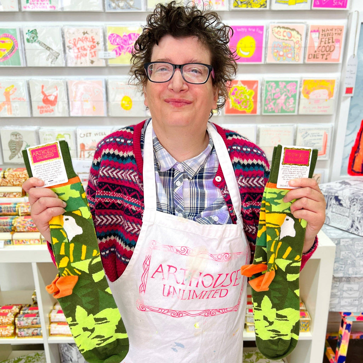 A person wearing glasses and a colorful knit sweater stands in a shop holding up a pair of green patterned socks with orange bows. The white apron on the person reads 'ARTHOUSE UNLIMITED'.