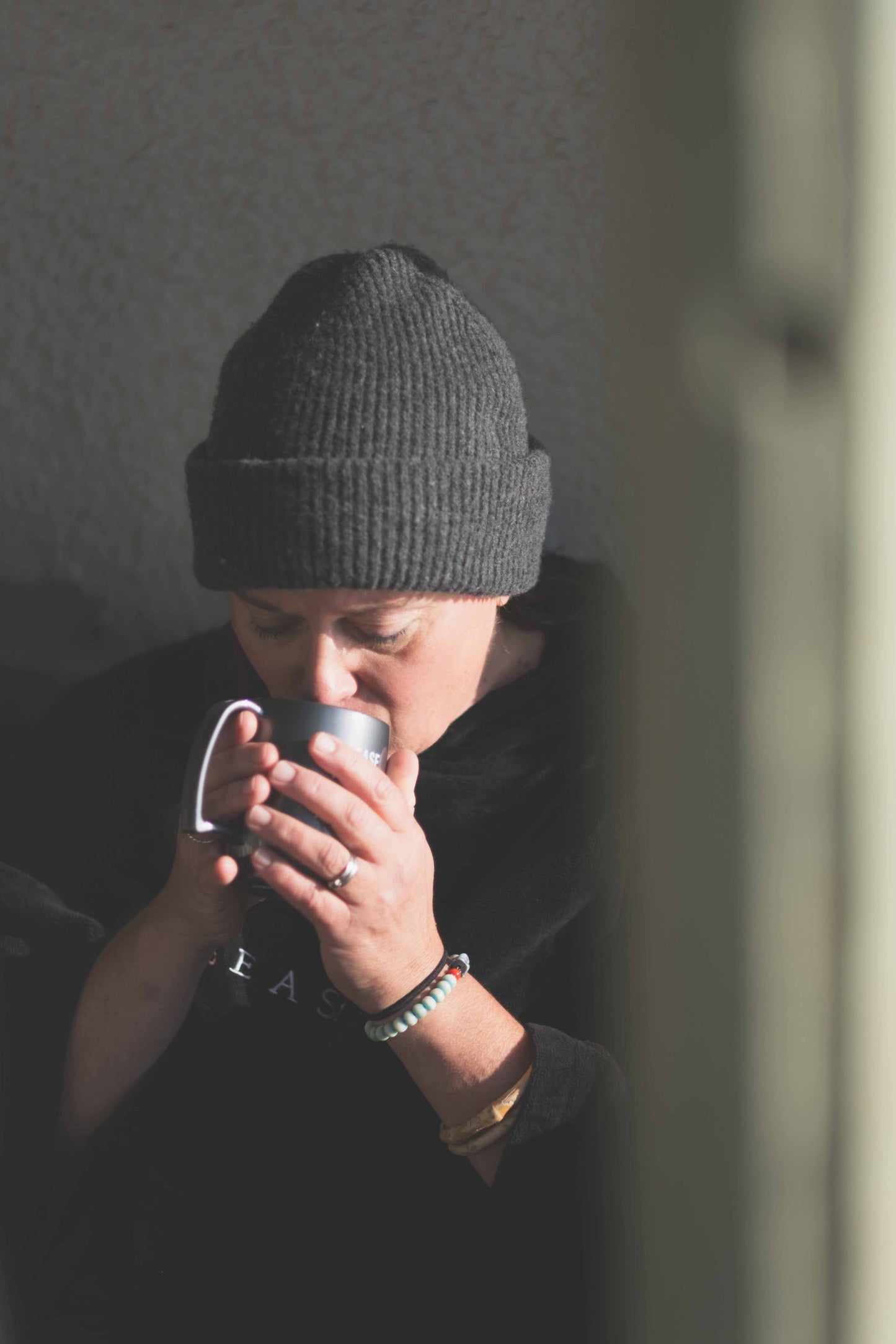 Person wearing a gray knit beanie and dark clothing, drinking from a mug.