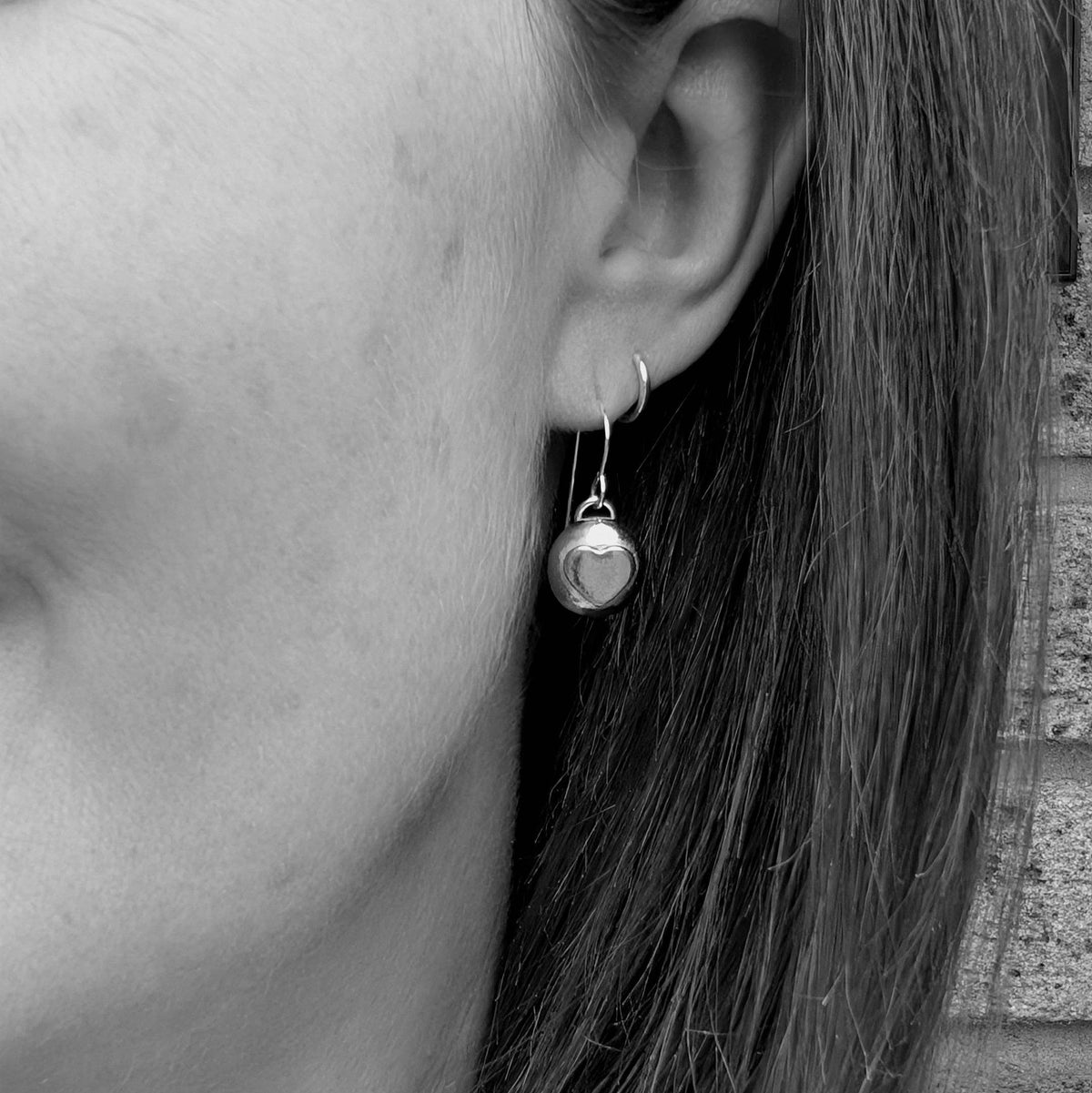 A close-up of a person's ear wearing a small silver hoop earring with a dangling polished silver heart charm.