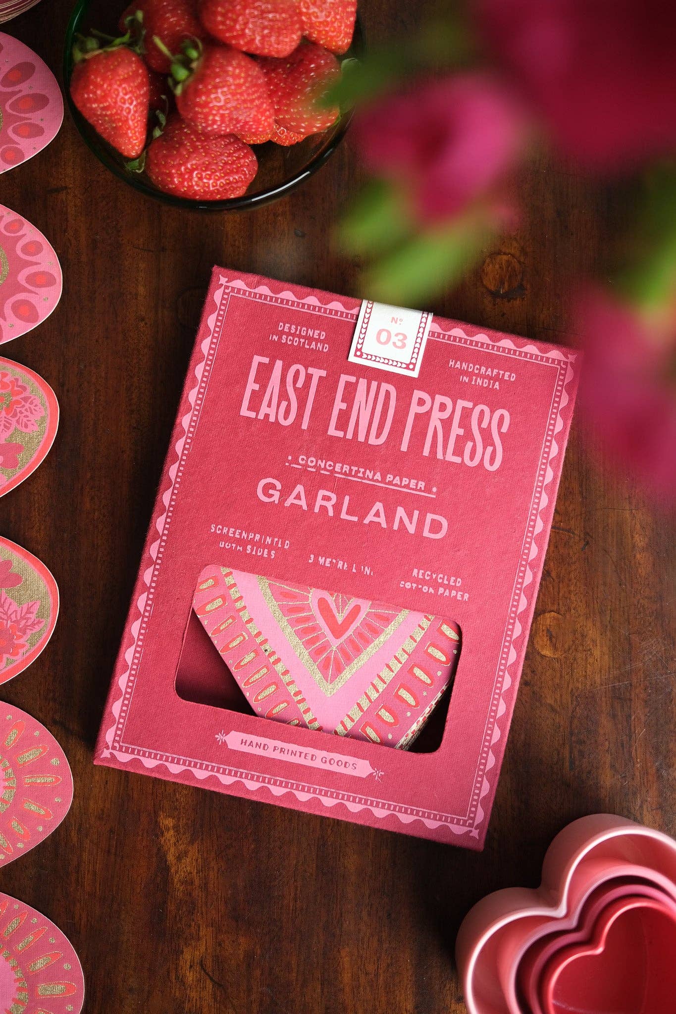 Pink packaging of 'Garland' by East End Press on a wooden surface with strawberries and flowers.