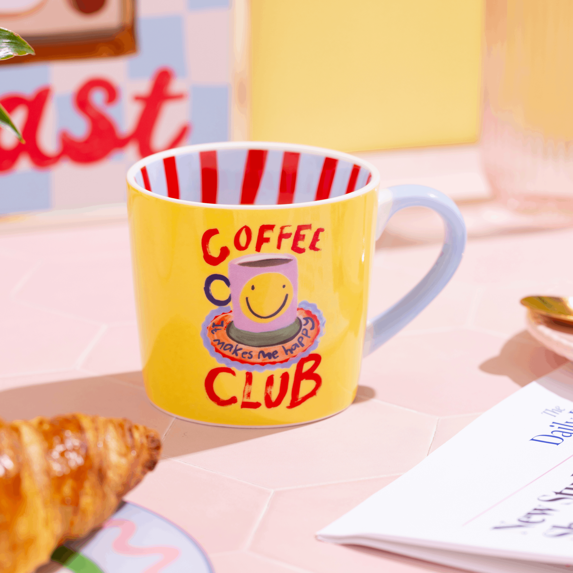 Yellow mug with red and white striped interior, 'Coffee Club' text, and smiley face design on a pink surface.