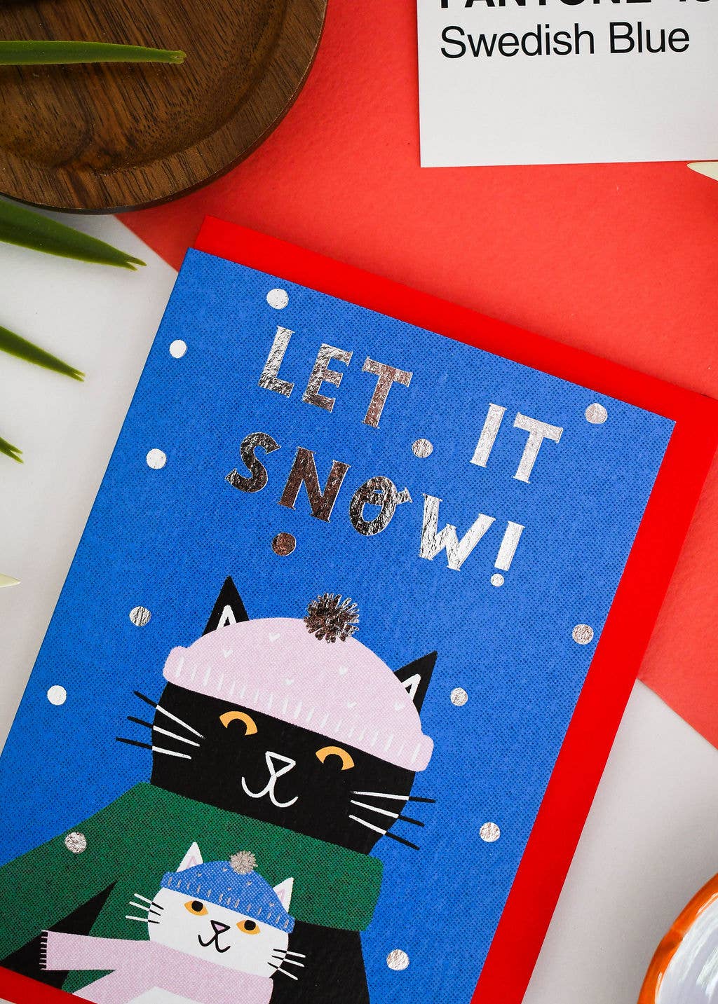 A festive blue greeting card with the words 'LET IT SNOW!' in metallic lettering, featuring two cats bundled in winter clothing, styled on a red envelope with a white card reading 'FANTASY Swedish Blue' in the background.