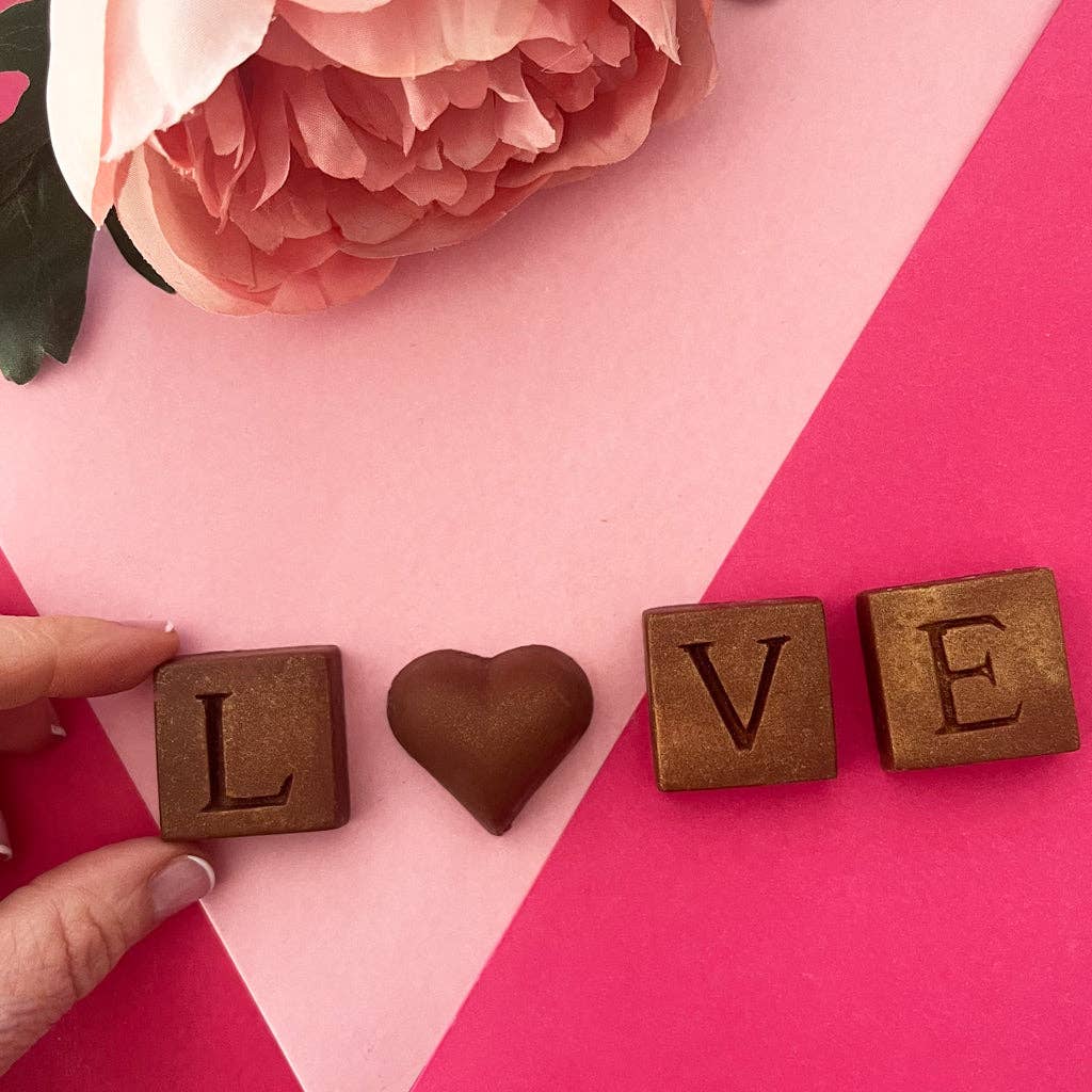 Wooden blocks spelling 'LOVE' with a chocolate heart on a pink background