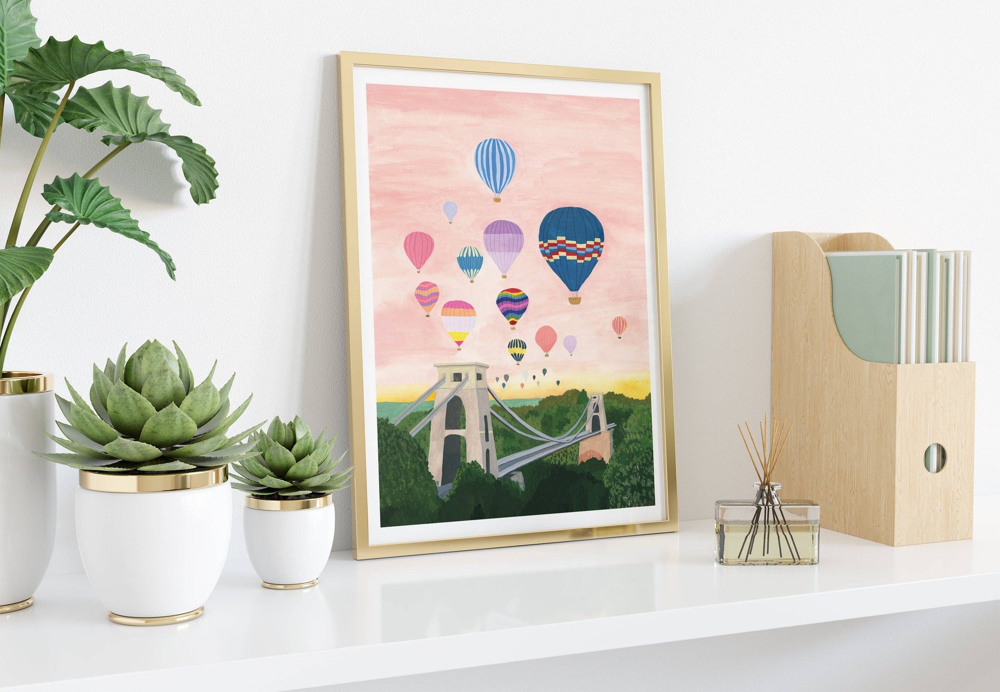Framed artwork of hot air balloons over a bridge on a white shelf with plants and a magazine holder.