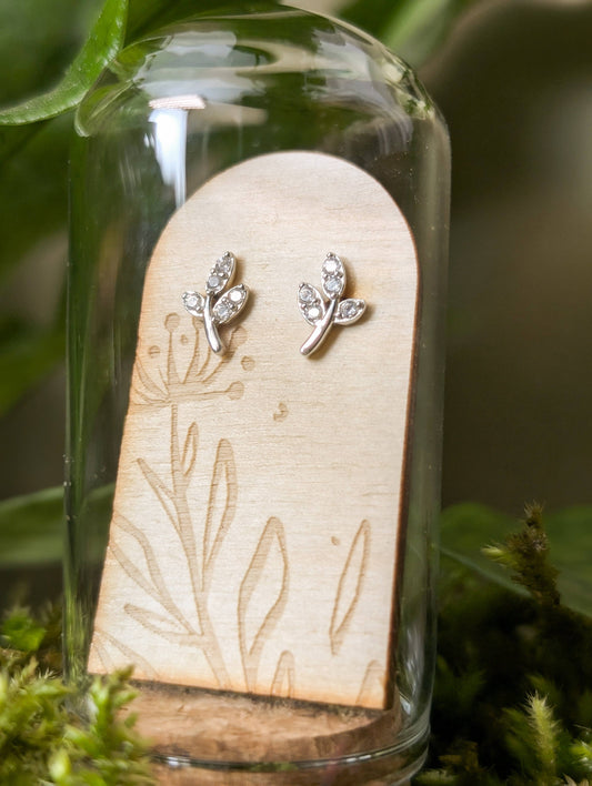 Pair of silver earrings on a wooden stand inside a glass dome with a natural background.