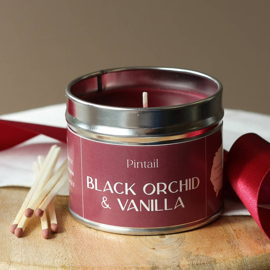 Candle in a tin labeled 'Black Orchid & Vanilla' by Pintail on a wooden surface with matches and a red ribbon.