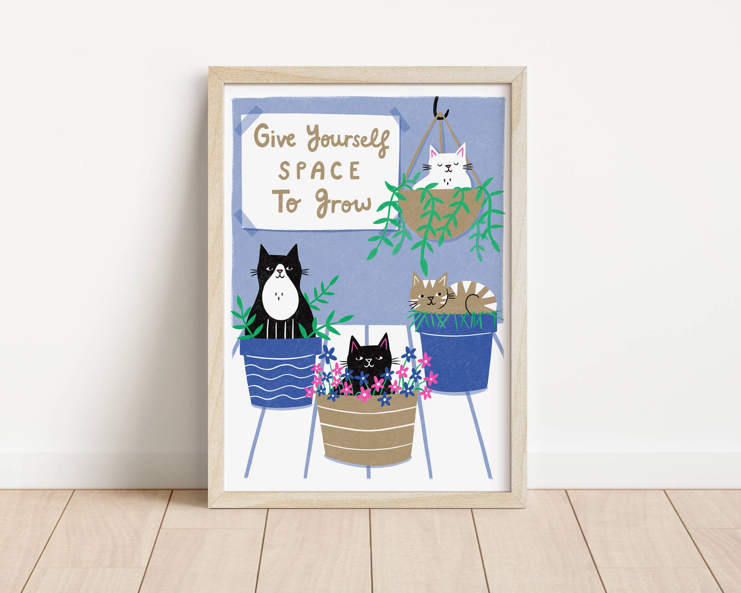 Framed artwork with cartoon cats in plant pots and motivational quote on a white wall.