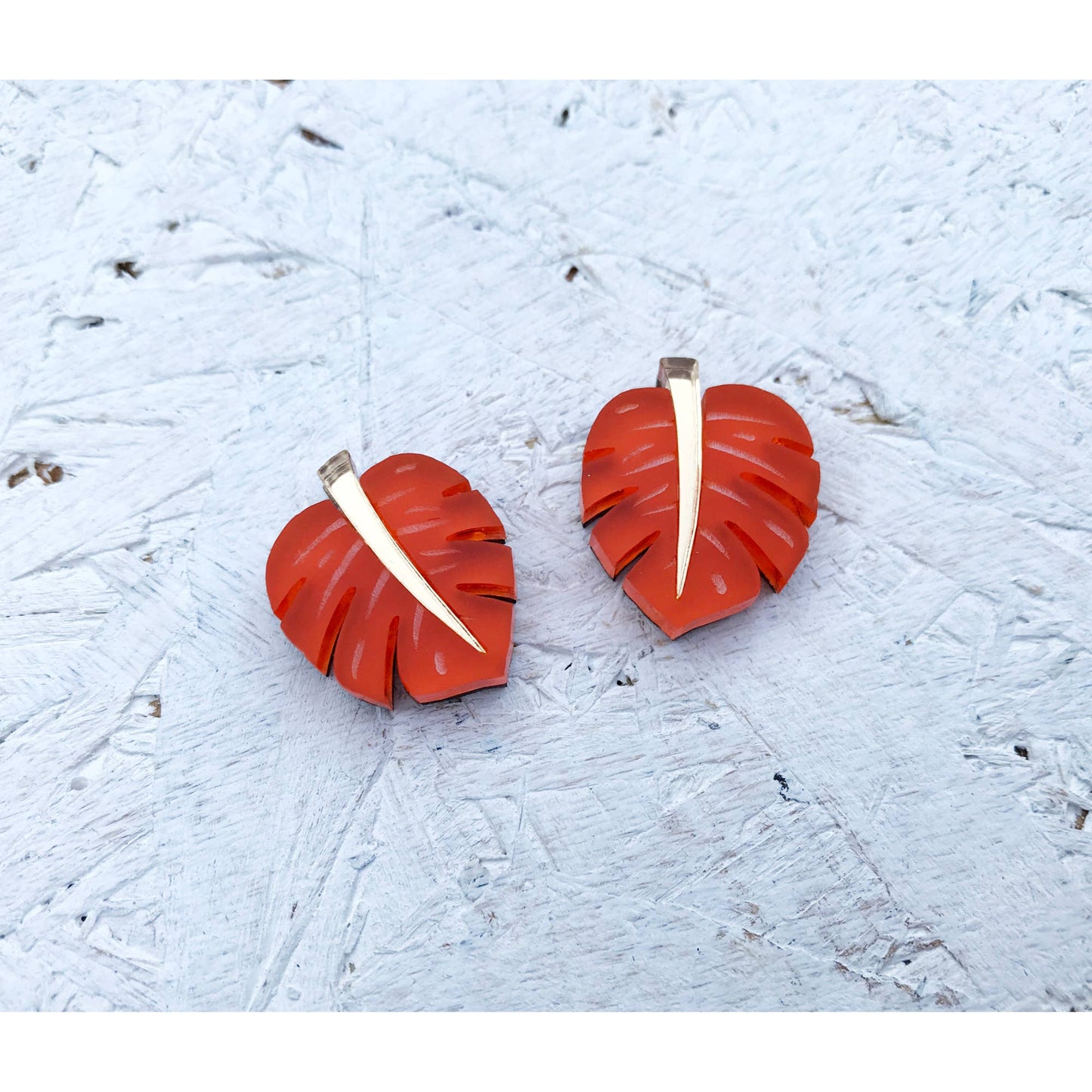 A pair of red monstera leaf stud earrings with a white center vein detail, displayed on a textured light gray surface.