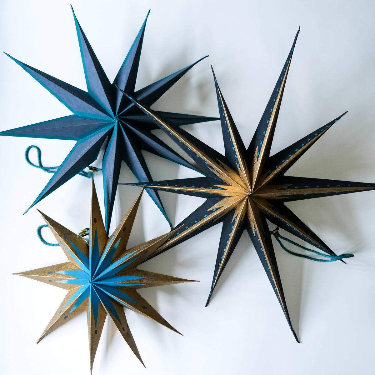 Three decorative star-shaped ornaments with geometric panel designs, displayed on a clean white background. The ornaments feature color combinations of black and teal, black and gold, and gold and teal, each with a small hanging string.
