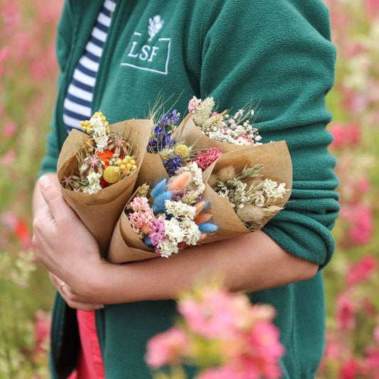 Person holding a bouquet of dried flowers wrapped in brown paper against a blurred floral background