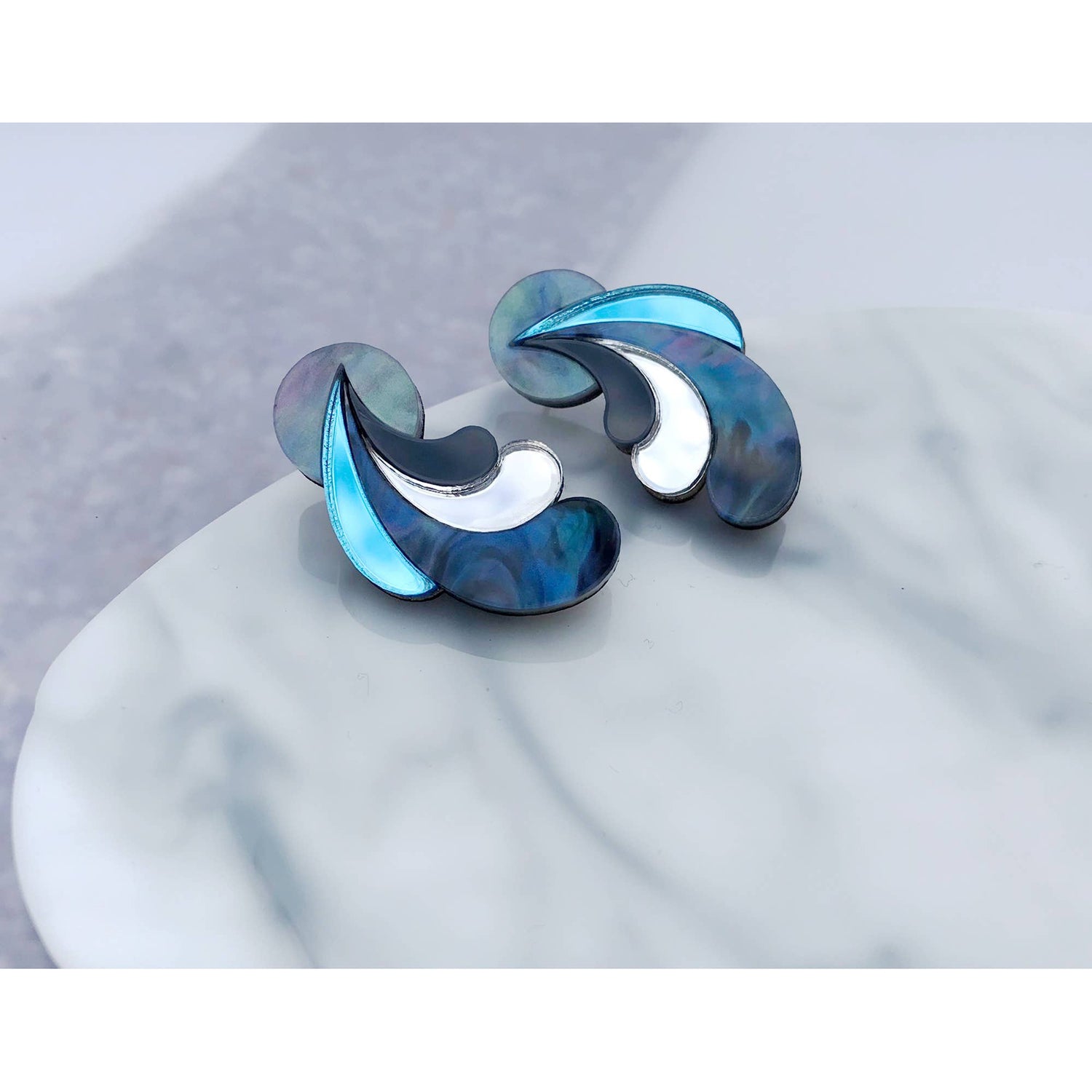 A pair of modern abstract wave-shaped earrings with iridescent blue, teal, and white abalone shell inlay, displayed on a white marble surface.