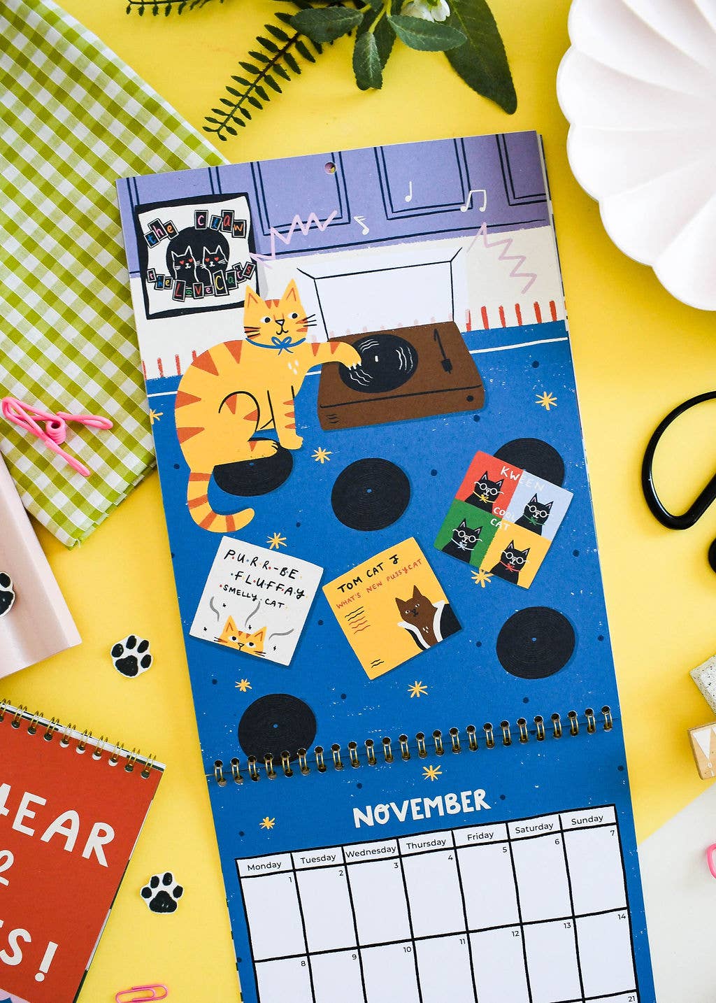 A spiral-bound November desk calendar featuring a whimsical cat-themed illustration on a blue background, styled in a flat-lay arrangement on a bright yellow surface with green gingham fabric, a red book, and small accessories.