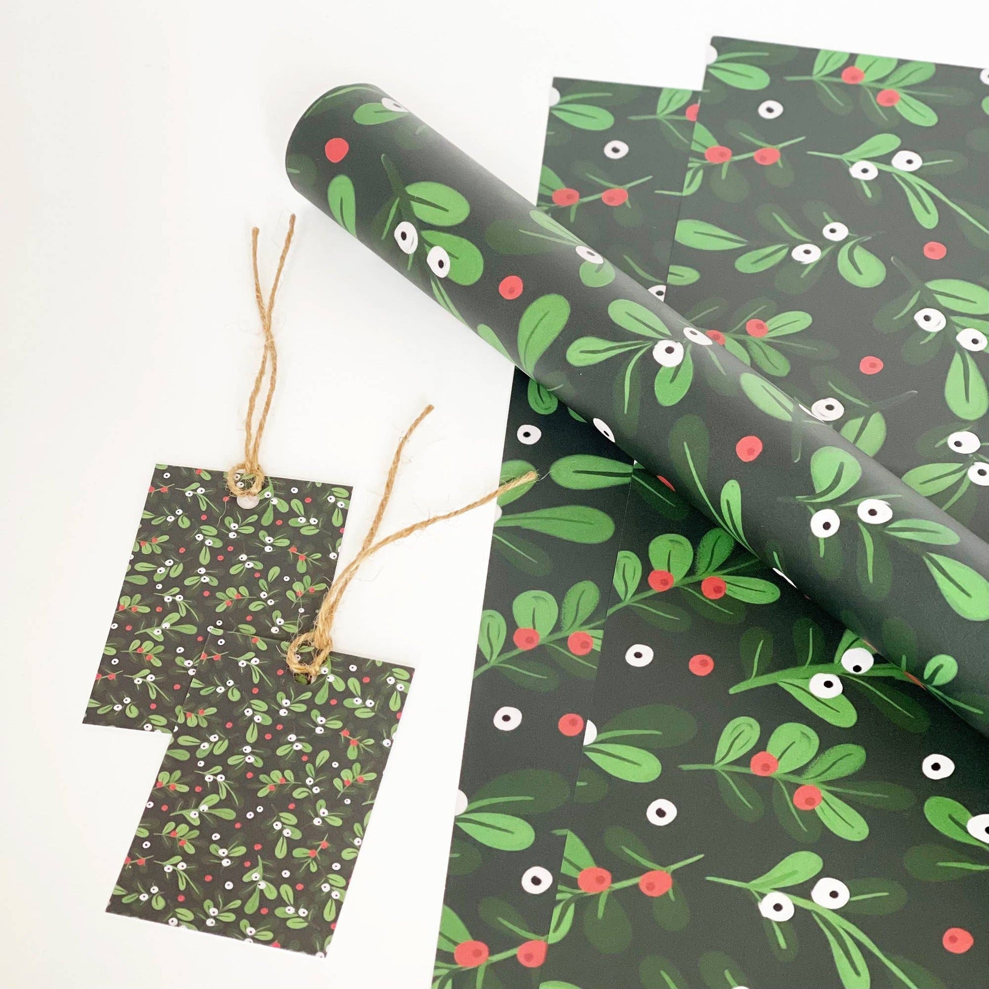 Holly and mistletoe patterned wrapping paper set with a rolled sheet and two matching gift tags on a white background.