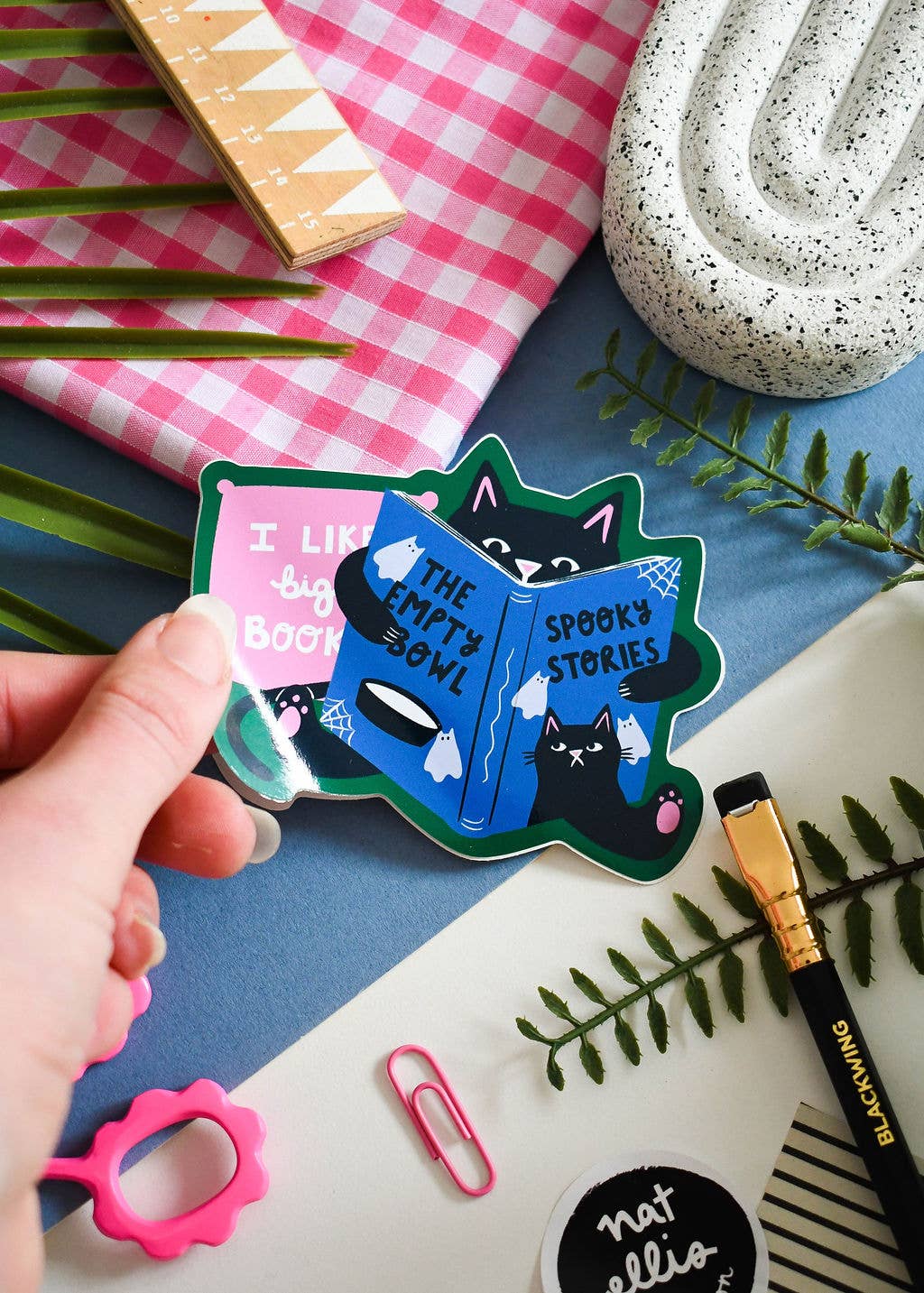 A hand holds a die-cut sticker featuring a spooky cat character reading a blue book titled 'The Empty Bowl' and black text reading 'Spooky Stories' and 'I like big books.' The scene is styled with a pink gingham cloth, green leaves, a white speckled bowl, a pink paperclip, and other stationery on a blue surface.