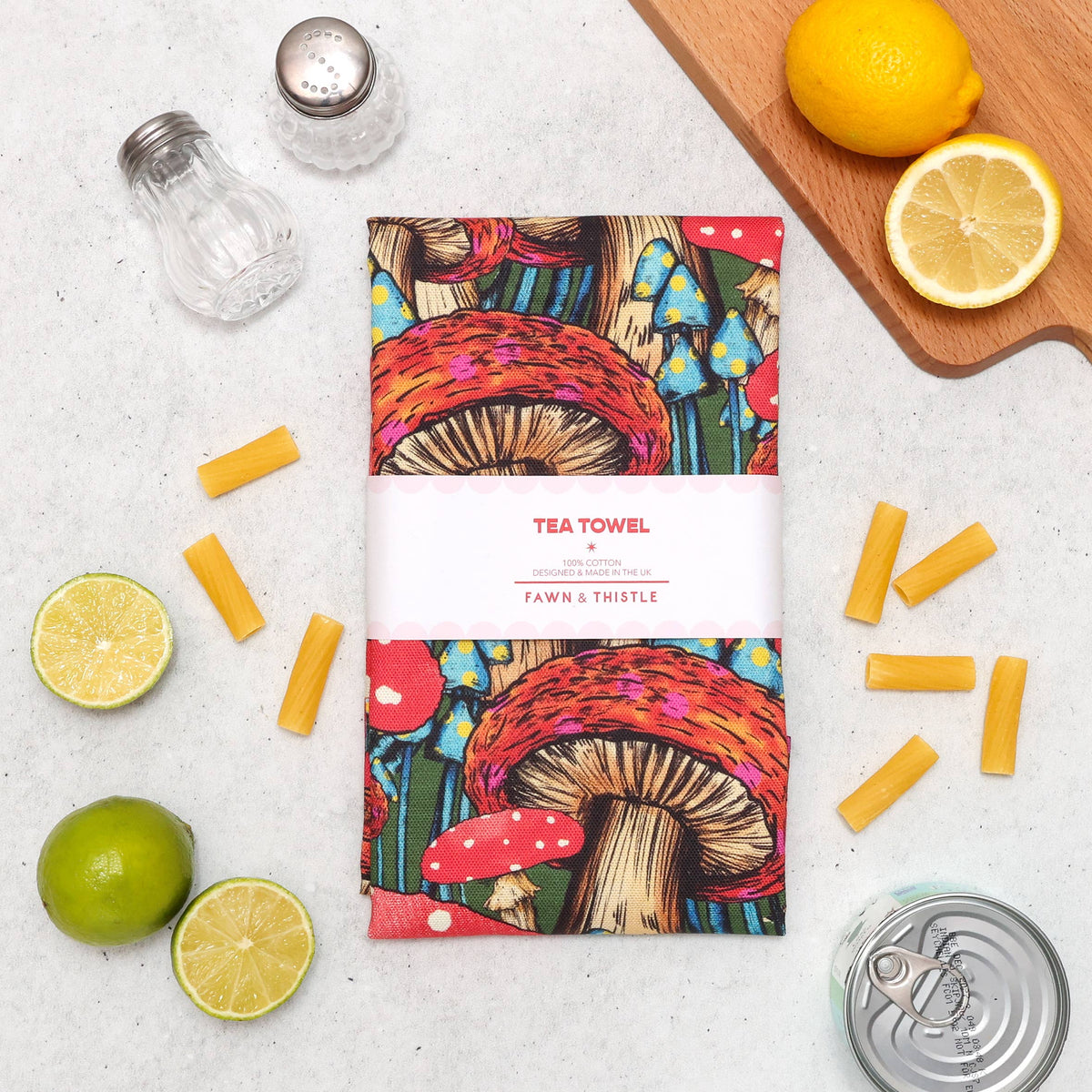 A printed tea towel with a colorful mushroom illustration and a white paper band reading 'TEA TOWEL' and 'FAWN & THISTLE' is arranged on a light surface with limes, pasta pieces, glass shakers, and a can opener around it.
