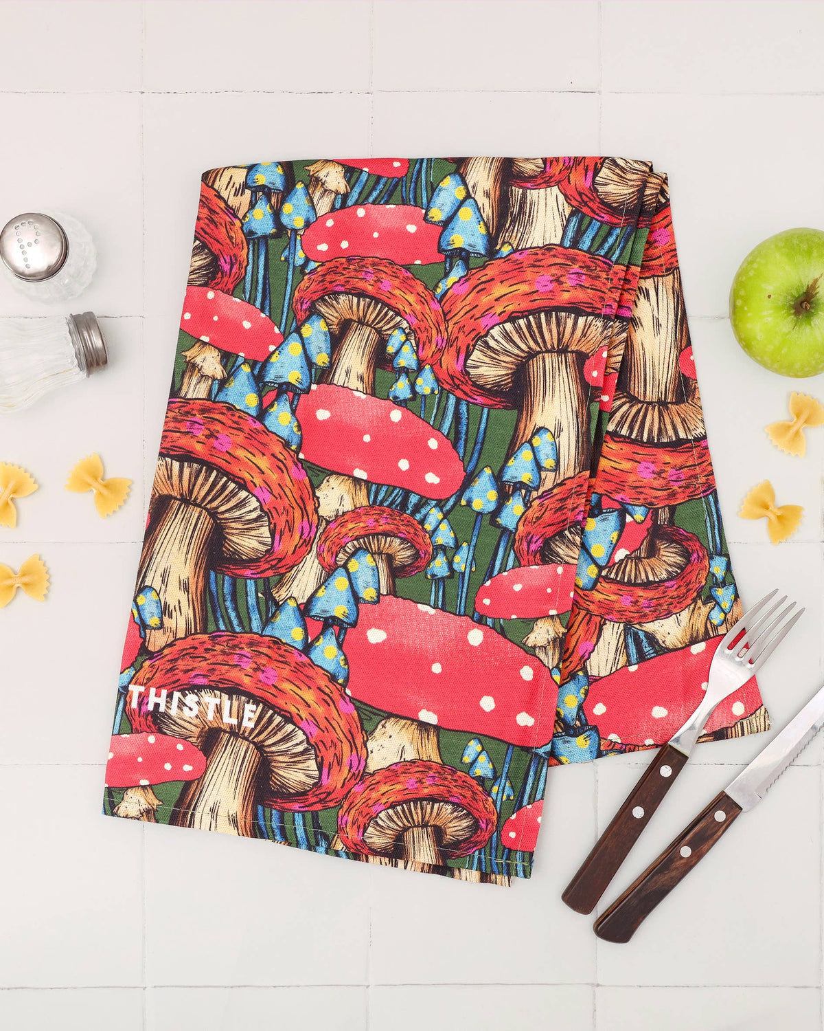 A kitchen towel with a colorful mushroom print, styled in a flat-lay arrangement on a white tiled background with a green apple, pasta, and cutlery.