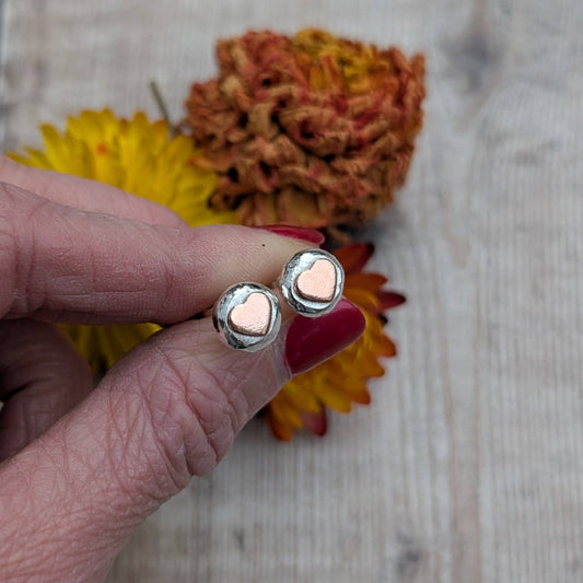 A close-up of a hand with red nail polish holding a pair of silver-toned stud earrings with small rose-gold heart-shaped centers, set against a rustic wooden background with dried yellow and orange flowers.