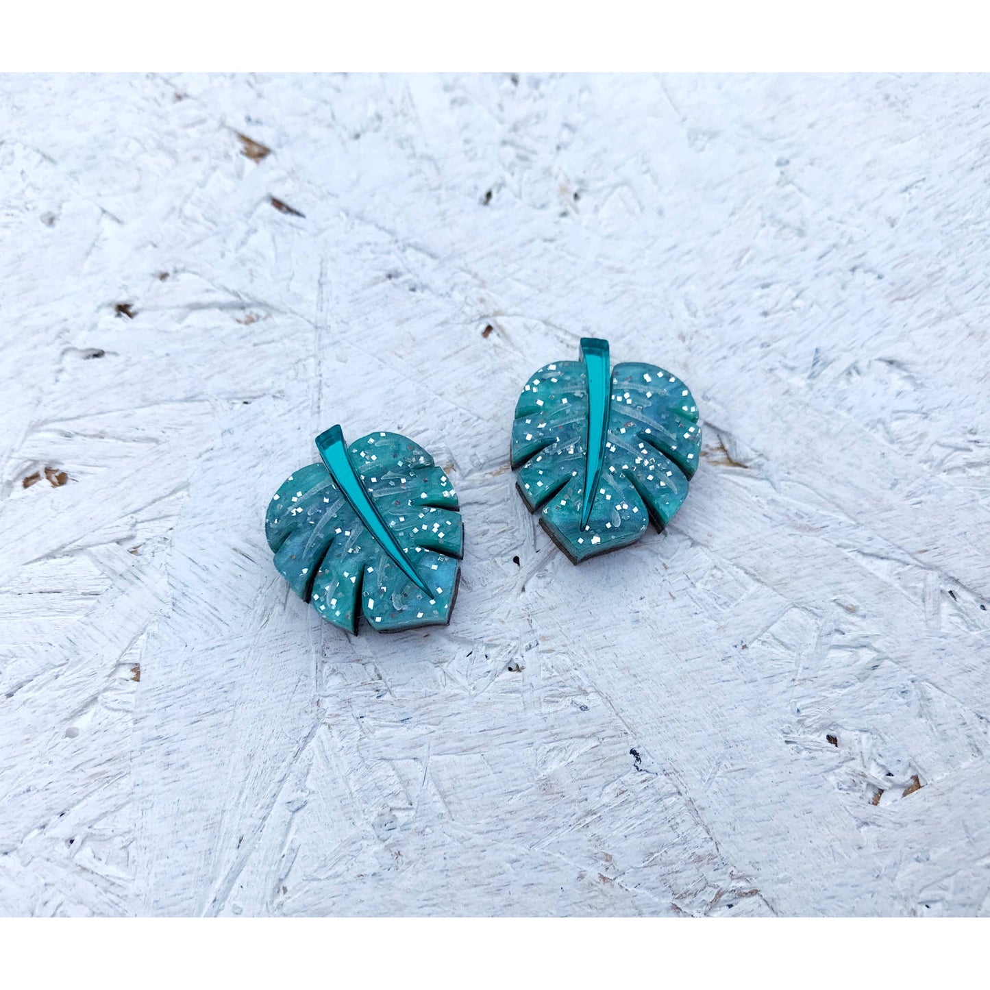 A pair of monstera leaf-shaped earrings with a teal and white speckled pattern and a central metallic teal bar, displayed on a textured white surface.