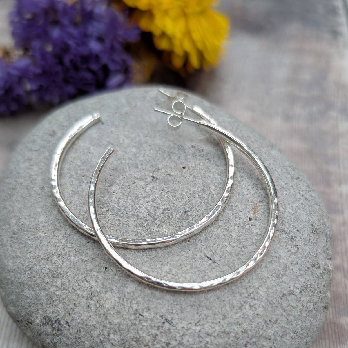 A pair of hammered silver hoop earrings resting on a smooth gray stone, with blurred purple and yellow flowers in the background.