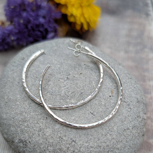 A pair of hammered silver hoop earrings resting on a smooth gray stone, with blurred purple and yellow flowers in the background.