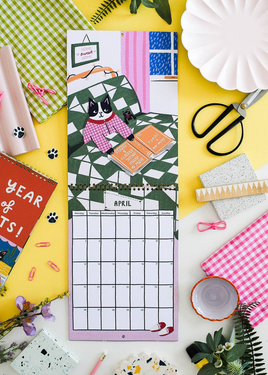 A decorative monthly calendar page with a cat illustration and a April grid, styled in a flat lay with scissors, paper plates, notebooks, and colorful stationery on a yellow background.