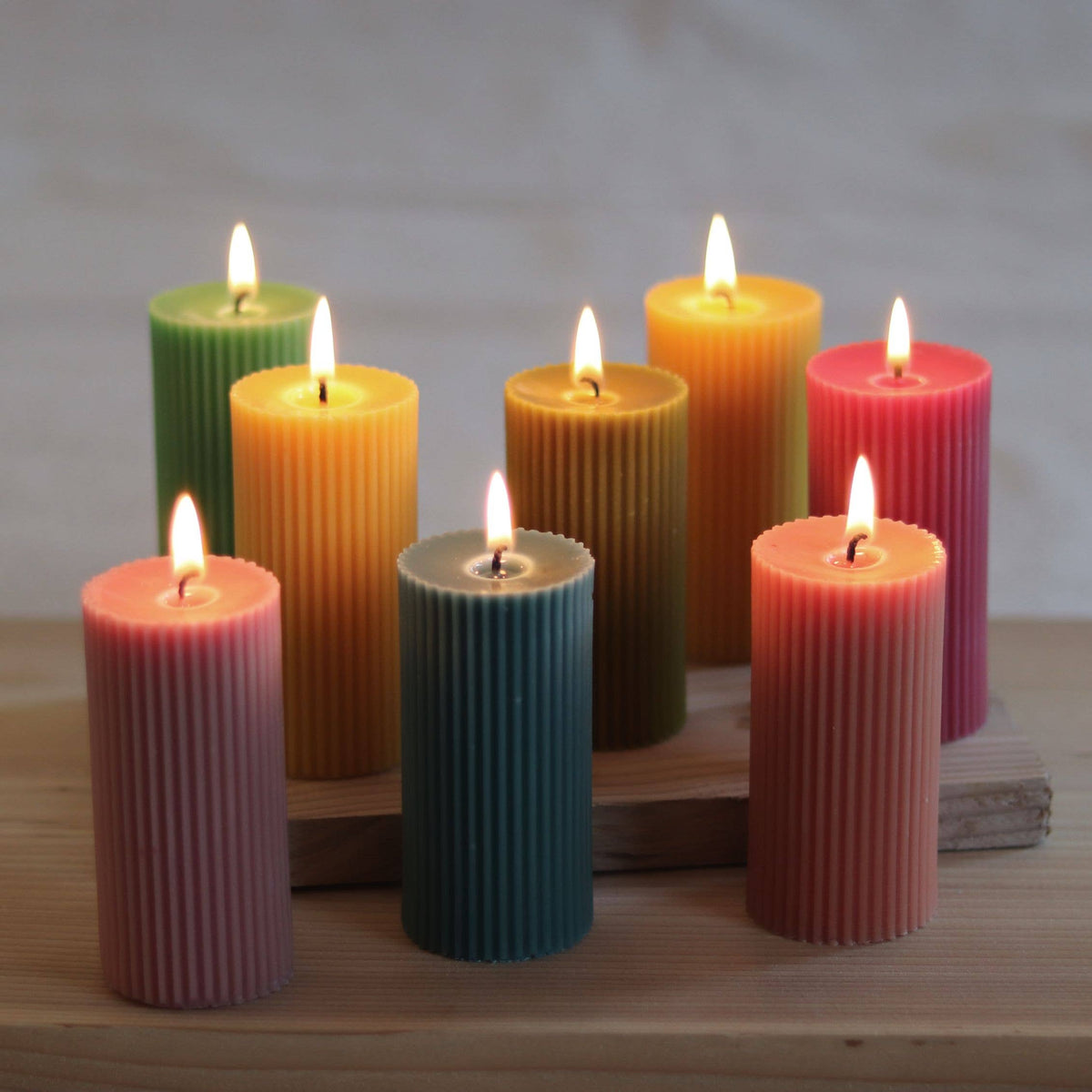 Six colorful ribbed pillar candles with lit flames, arranged on a wooden block against a soft neutral background.