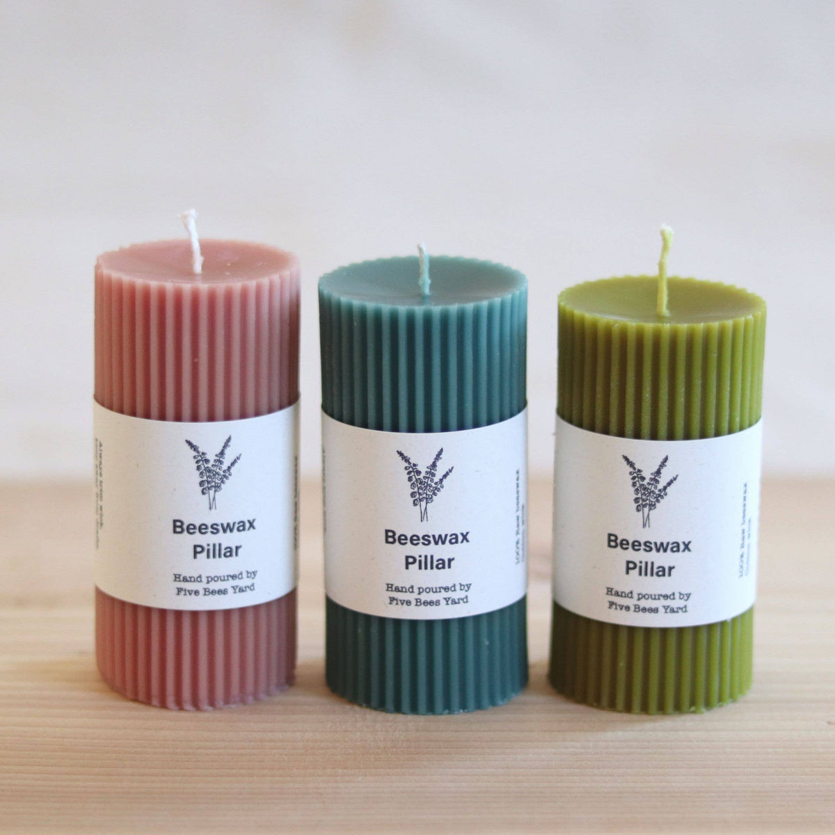Three ribbed pillar candles in dusty pink, teal, and olive green, each wrapped with a white paper label reading 'Beeswax Pillar' and 'Hand poured by Five Bee Tinct.' on a light wood surface against a soft neutral background.